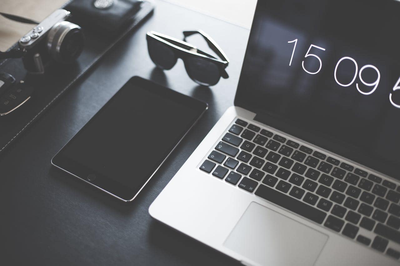 A flat lay of a modern workspace featuring a laptop, tablet, camera, and sunglasses.